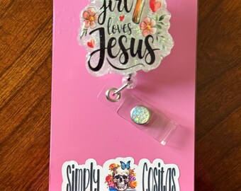 May include: A pink badge holder with the text "This girl loves Jesus" and a cross design. The holder also features the text "Simply Cositas" and a skull with a butterfly and flowers.