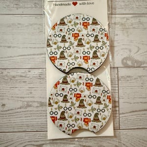 May include: Two car coasters with a white background and a repeating pattern of a sorting hat, glasses, and letters. The coasters are packaged in a clear plastic sleeve. The text "Car Coasters" and "Handmade with love" are printed on the packaging.