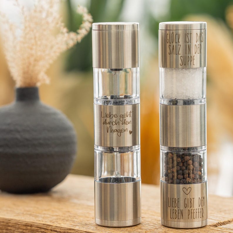May include: Two stainless steel salt and pepper grinders with clear glass containers. The salt grinder has the text "Gl&uuml;ck ist das Salz in der Suppe" and the pepper grinder has the text "Liebe geht durch den Magen" and "Liebe gibt dem Leben Pfeffer".