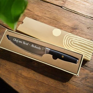 Könnte beinhalten: Ein silbernes Kochmesser mit schwarzem Griff in einer braunen Box. Das Messer ist mit dem Text "Chef am Herd - Michaela" und einem Herz graviert.