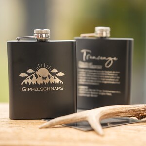 Könnte beinhalten: Zwei schwarze Metall-Hüftflaschen mit gravierten Designs. Eine Flasche zeigt eine Bergszene mit einer Sonne und dem Text "GIPFELSCHNAPS". Die andere Flasche hat das Wort "Trinkauge" eingraviert.