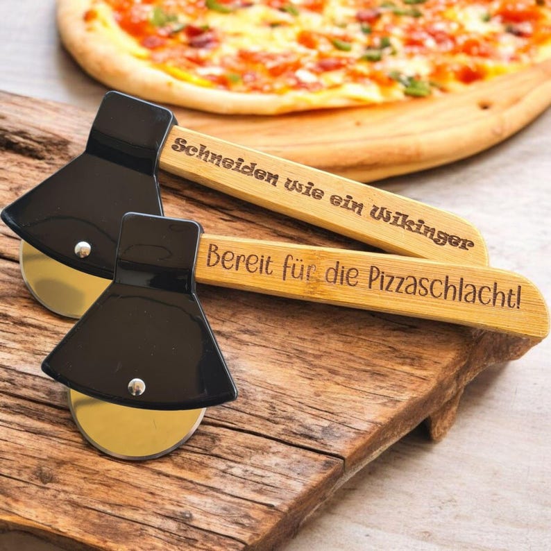 May include: Two wooden pizza cutters shaped like axes with black metal blades. The handles are engraved with the text "Schneiden wie ein Wikinger" and "Bereit f&uuml;r die Pizzaschlacht!"