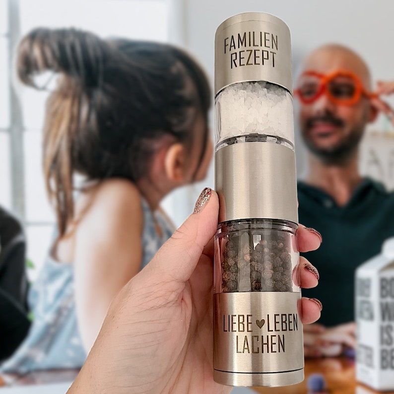 May include: A silver salt and pepper grinder with the text "Familien Rezept" on the top section, "Liebe Leben Lachen" on the bottom section, and salt and peppercorns visible inside.
