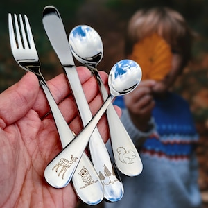 Könnte beinhalten: Ein Set aus Edelstahlbesteck, einschließlich einer Gabel, eines Messers und zwei Löffeln. Die Löffel haben reflektierende Oberflächen. Die Löffelgriffe zeigen eingravierte Bilder eines Hirsches, einer Eule, Bäumen und eines Eichhörnchens. Das Besteck wird in einer Hand gehalten.