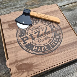 May include: A wooden pizza box with a black pizza cutter shaped like an axe. The box has a brown woodgrain pattern and the text "Finest Pizza Quality Made Fresh" in a black circle. The pizza cutter has a wooden handle with the text "Bereit fur die Pizzaschlacht!" engraved on it.