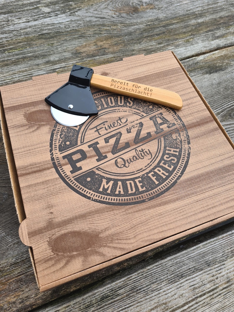 May include: A wooden pizza box with a black pizza cutter shaped like an axe. The box has a brown woodgrain pattern and the text "Finest Pizza Quality Made Fresh" in a black circle. The pizza cutter has a wooden handle with the text "Bereit fur die Pizzaschlacht!" engraved on it.