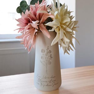 Könnte beinhalten: Eine beige Keramikvase mit floralem Design und Text, die rosa und cremefarbene Kunstblumen enthält. Die Vase hat eine leicht konische Form und steht auf einer Holzoberfläche. Der Text auf der Vase lautet "Ein Leben ohne Blumen ist wie ein Tag ohne Sonne."