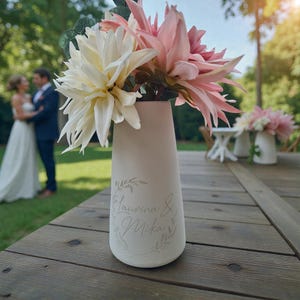 Könnte beinhalten: Eine cremefarbene Keramikvase mit weißen und rosa Dahlien. Die Vase ist mit den Namen "Laurina & Mika" in einem Kranz graviert. Die Vase steht auf einem Holztisch, im Hintergrund ein Hochzeitspaar.
