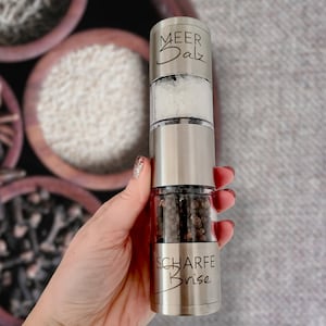 May include: A silver salt and pepper grinder with a clear top. The salt grinder is labeled "Meer Salz" and the pepper grinder is labeled "Scharfe Brise".