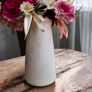Könnte beinhalten: Weiße Keramikvase mit floralem Design und dem Text "ES IST SCHÖN DASS ES DICH GIBT" in einer kursivschriftlichen Schriftart. Die Vase ist mit rosa und weißen Blumen gefüllt.