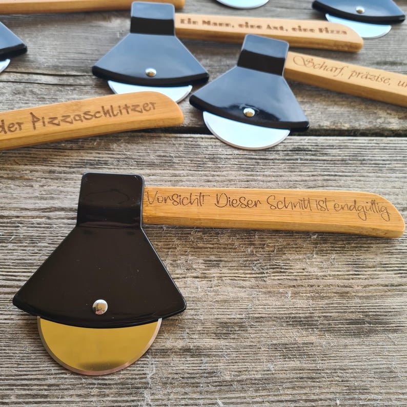 May include: A black pizza cutter with a wooden handle. The handle is engraved with the text "Vorsicht Dieser Schnitt ist endgillig".