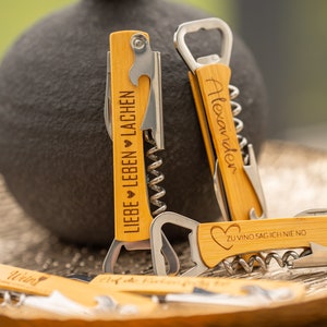 Könnte beinhalten: Vier Flaschenöffner aus Holz mit verschiedenen gravierten Sprüchen. Die Sprüche lauten "Liebe Leben Lachen", "Alexander", "Zu Vino Sag Ich Nie No" und "Wein".