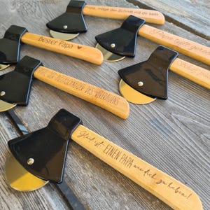 May include: Set of five wooden pizza cutters shaped like axes with black handles and engraved messages. The messages include "Best Papa", "Papa teilt gerne", "Scharf, pras", "Dir PIZZASCHNEIDER DES VERTRAUENS", and "Gl&uuml;ck ist EINEN PAPA wie dich zu haben!"