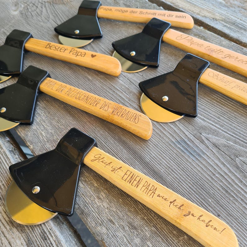 May include: Set of five wooden pizza cutters shaped like axes with black handles and engraved messages. The messages include "Best Papa", "Papa teilt gerne", "Scharf, pras", "Dir PIZZASCHNEIDER DES VERTRAUENS", and "Gl&uuml;ck ist EINEN PAPA wie dich zu haben!"
