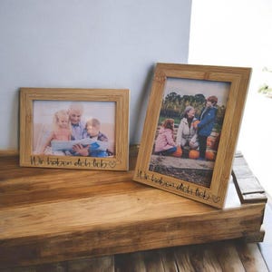 Könnte beinhalten: Zwei rechteckige Holzbilderrahmen mit Familienfotos. Die Rahmen haben eine natürliche Holzoberfläche und der Text "Wir haben dich lieb" ist unten eingraviert. Ein Rahmen steht aufrecht, der andere liegt flach.