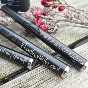 Könnte beinhalten: Drei schwarze Feuerzeuge mit silbernen Akzenten und Schriftzügen. Die Feuerzeuge liegen auf einer Holzoberfläche mit dekorativen Elementen. Der Text ist in einer Schreibschrift. Die Feuerzeuge haben ein schlankes, modernes Design.