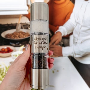 May include: A silver salt and pepper grinder with a clear glass section. The grinder has an inscription that reads "Liebe geht durch den Magen" with a small heart symbol.