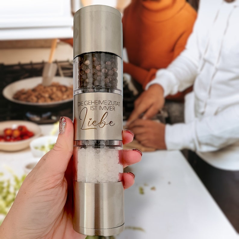 May include: A silver salt and pepper grinder with a black peppercorn and white salt filling. The grinder has the text "Die geheime Zutat ist immer Liebe" engraved on the side.