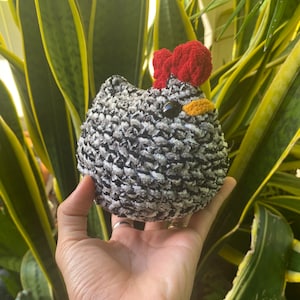 May include: A black and white crocheted chicken plush toy with a red bow on its head. The toy has a yellow beak and black eyes.