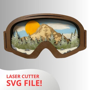 May include: A laser cut wooden design of a mountain scene viewed through a pair of ski goggles. The scene includes a yellow sun, a brown mountain, green trees, and white snow. The text "LASER CUTTER SVG FILE!" is printed below the goggles.