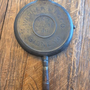May include: A round, dark grey cast iron waffle iron plate with a wooden handle. The surface features embossed text reading WAGNER MFG. CO., PATD JULY 26 1892, and SIDNEY O. The metal has a weathered, dark patina and a circular hinge component at the top.