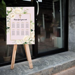 May include: A wedding seating plan displayed on a wooden easel. The sign is pale pink with a gold border and features a cross and the text "Please find your seat." White roses and foliage decorate the edges.