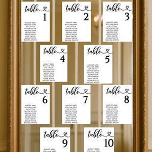 May include: A gold framed mirror displays ten white table assignment cards. Each card has a table number and a list of names in black script. The word "table" is written above each number with a heart symbol.