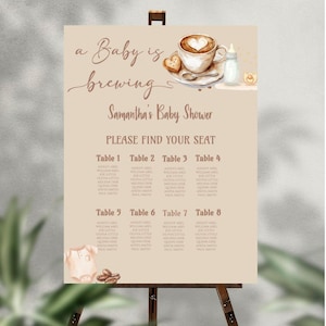 May include: A baby shower seating chart on a wooden easel. The sign is beige with the text "a Baby is brewing" and "Samantha's Baby Shower." It includes a coffee cup, cookie, and baby bottle illustration. Tables 1-8 list guest names.