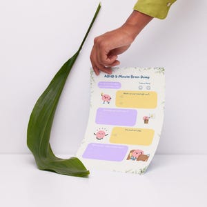 Può includere: Un foglio di carta bianco con il testo "ADHD 5-Minute Brain Dump" e caselle colorate per scrivere. Il foglio è tenuto da una mano, con una grande foglia verde in primo piano.
