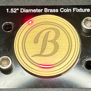 May include: A 1.52" diameter brass coin fixture with a circular cutout and a letter "B" engraved in the center. The fixture is mounted on a black surface with four screw holes.