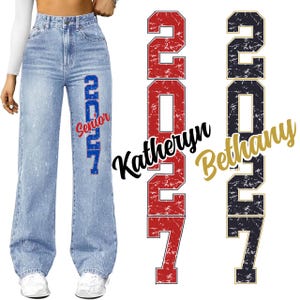 Può includere: Jeans di denim chiaro con gamba larga. I jeans presentano la scritta "Senior 2027" in blu e rosso. Testo e numeri aggiuntivi visibili in rosso, nero e oro.