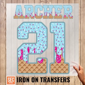May include: An iron-on transfer featuring the name "ARCHER" above the number "21". The text and numbers are light blue with pink sprinkles and dripping pink icing. The bottom of the numbers have a waffle cone design. The words "IRON ON TRANSFERS" are below.