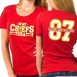 Puede incluir: Camiseta roja con el texto "IN MY CHEFS ERA" en blanco y amarillo en la parte delantera. La parte trasera de la camiseta muestra el número "87" en blanco y amarillo, con la frase "LOVING HIM WAS RED" debajo.