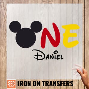 May include: Iron-on transfer featuring a black Mickey Mouse head, the word "ONE" in red and yellow, and the name "Daniel" in black script. The design is on a clear sheet with the text "IRON ON TRANSFERS".
