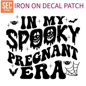 May include: Black iron-on decal patch with the text "IN MY SPOOKY PREGNANT ERA" in a retro font. The design includes Halloween-themed graphics such as spiders, bats, and a spider web. The patch also has the text "SEC APPAREL".