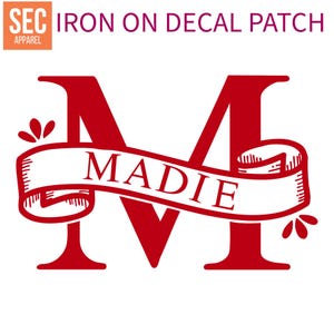 以下が含まれることがあります： 赤いアイロン接着デカールパッチ。大きな文字「M」と「Madie」という名前がバナーに書かれています。パッチには、バナーの両側に装飾的な花の要素があります。「Iron on decal patch」というテキストが文字の上にあります。