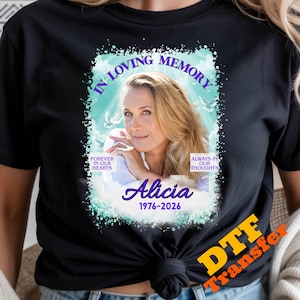 May include: A black t-shirt featuring a memorial graphic with a portrait of a woman, the text IN LOVING MEMORY, FOREVER IN OUR HEARTS, ALWAYS IN OUR THOUGHTS, Alicia, and the dates 1976-2026, set against a light blue sky background with white clouds.