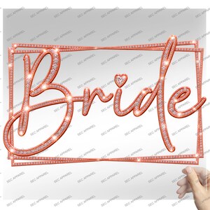May include: A rose gold-toned decorative sign featuring the word Bride in a cursive script font, accented with a small heart above the letter i. The entire design, including the rectangular border, is encrusted with numerous small, sparkling crystals.