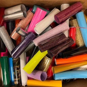 Puede incluir: Una caja llena de rollos de vinilo de purpurina de colores, incluyendo rosa, morado, azul, verde, dorado y rojo. El vinilo se utiliza para manualidades y decoración.