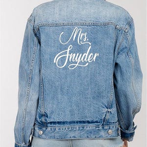 May include: A light blue denim jacket with a white embroidered design that says "Mrs. Snyder" on the back.
