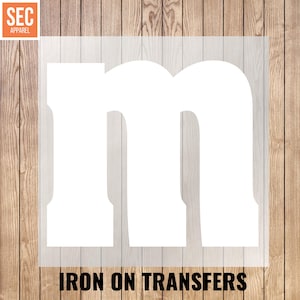 Puede incluir: Transferencia de planchado blanca con la letra "m" sobre una lámina transparente. El texto "IRON ON TRANSFERS" está impreso en negro en la parte inferior. El fondo es una superficie de madera con tablones verticales. El logotipo de SEC Apparel está en la esquina superior izquierda.