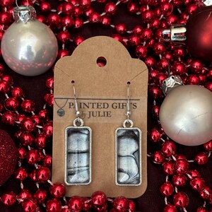 May include: A pair of silver-toned rectangular earrings with a marbled black and silver design. The earrings are displayed on a brown tag with the text "PAINTED GIFTS by JULIE". The background features red beads and Christmas ornaments.