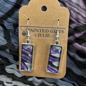 May include: Silver-toned rectangular earrings with a purple, gray, and black abstract design. The earrings hang from silver hooks and are displayed on a brown card with the text "PAINTED GIFTS by JULIE". The background is a patterned fabric.