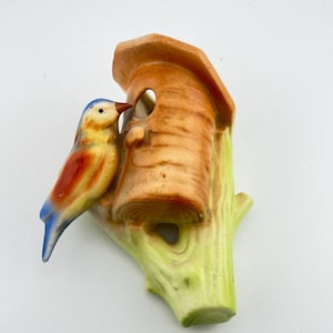 May include: A ceramic wall ornament featuring a small bird with a blue head, yellow chest, and orange wings perched on a light green tree branch next to a brown, cylindrical birdhouse with a circular opening and a sloped roof.