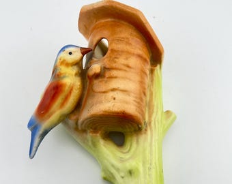May include: A ceramic wall ornament featuring a small bird with a blue head, yellow chest, and orange wings perched on a light green tree branch next to a brown, cylindrical birdhouse with a circular opening and a sloped roof.