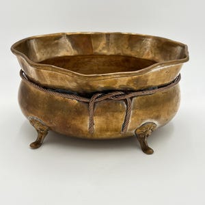 May include: A round brass bowl with a hammered texture and a wavy rim. The bowl features three ornate, claw-style feet and is encircled by a decorative twisted copper-colored wire rope detail tied in a knot at the front center.