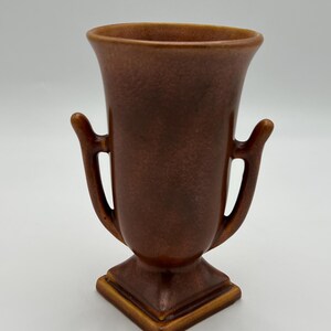 May include: A brown ceramic vase with a flared rim, two side handles, and a square base. The surface features a mottled, matte finish with subtle variations in tone, creating a textured appearance across the body of the vessel.
