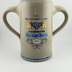 May include: A beige ceramic beer stein with two handles, featuring blue horizontal bands at the top and bottom. The front displays the text "Mit beiden Händen greifen...", "KULMBACHER Reichelbräu", and "Sammlerkrug Der Doppelhänder" with a crest.