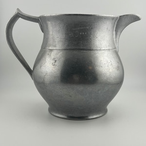 May include: A vintage pewter pitcher with a handle and spout. The pitcher has a bulbous body and a slightly textured surface. The handle is curved, and the spout is pointed. The pitcher is a muted gray color and appears to be antique.