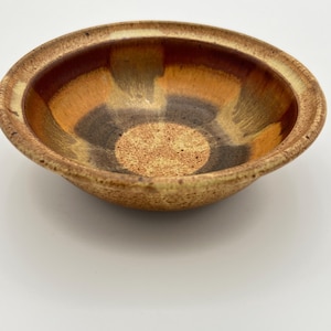 May include: A round ceramic bowl featuring a speckled tan exterior and a smooth interior glaze. The interior displays a gradient of colors, transitioning from a textured tan center to dark brown and golden yellow bands toward the rim.